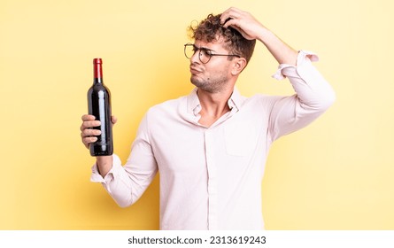 young handsome man feeling puzzled and confused, scratching head. wine bottle concept