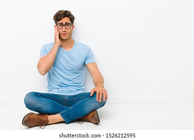 young handsome man feeling shocked and astonished holding face to hand in disbelief with mouth wide open sitting on the floor