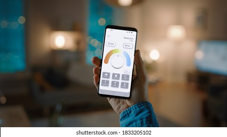 Young Handsome Man Gives a Voice Command to a Smart Home Application on His Smartphone To Set a Comfortable Light Temperature. It's a Cozy Evening in Technologically Advanced Living Room. - Powered by Shutterstock