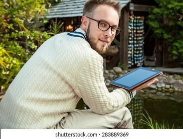 Young Handsome Man In Glasses Sit In Luxury Sofa With IPad In Summer Garden. 