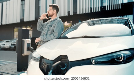 Young handsome man having conversation on smartphone while waiting till his electric car charge. Male standing on street chatting on cellphone while charging electric vehicle. Eco transport, future - Powered by Shutterstock