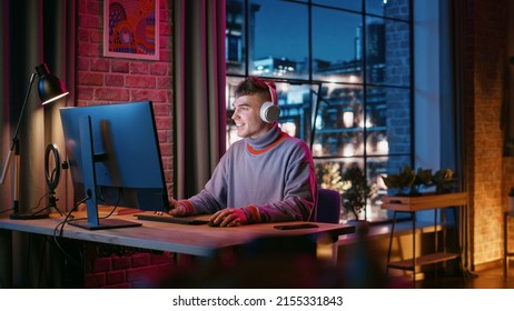 Young Handsome Man in Headphones Using Computer in Stylish Loft Apartment at Night. Creative Male Working from Home, Browsing Internet and Social Media. Urban City View from Big Window. - Powered by Shutterstock