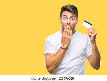 Young handsome man holding credit card over isolated background cover mouth with hand shocked with shame for mistake, expression of fear, scared in silence, secret concept