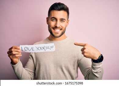 Young Handsome Man Holding Paper With Self Love Message Over Pink Background With Surprise Face Pointing Finger To Himself