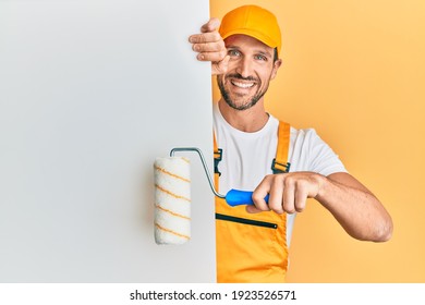 Young handsome man holding roller painter over white banner smiling with a happy and cool smile on face. showing teeth. 