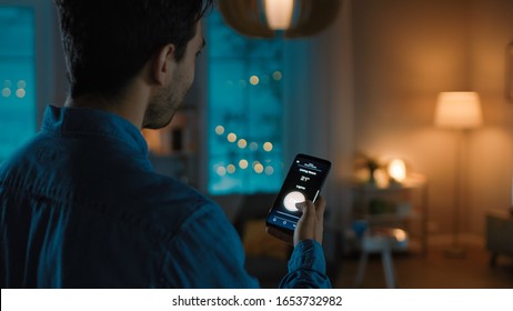 Young Handsome Man is Holding Smartphone with Active Smart Home Application. Person is Tapping the Screen to Turn On/Off the Lights in the Room. It's Cozy Evening in the Apartment.