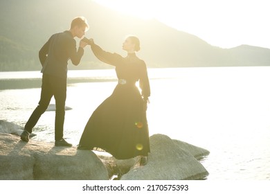 Young Handsome Man Kisses Hand Of A Beautiful Young Woman Standing On The Seacoast At Sunset. Historical Romance Novel. Loving  Relationship. 19th Century Style.