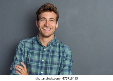 Young Handsome Man Leaning Against Grey Wall With Arms Crossed. Cheerful Man Laughing And Looking At Camera With A Big Grin. Portrait Of A Happy Young Man Standing Over Grey Background.