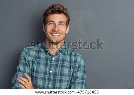 Young handsome man leaning against grey wall with arms crossed. Cheerful man laughing and looking at camera with a big grin. Portrait of a happy young man standing over grey background.