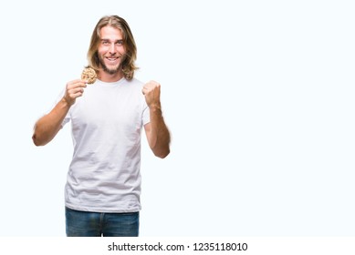 Young handsome man with long hair eating chocolate cooky over isolated background screaming proud and celebrating victory and success very excited, cheering emotion