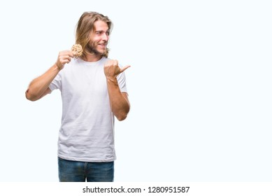 Young handsome man with long hair eating chocolate cooky over isolated background pointing and showing with thumb up to the side with happy face smiling