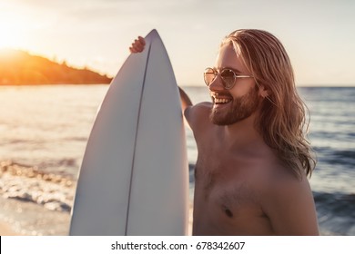 Young handsome man with long hair is standing on beach with white surfboard in hands and smiling. - Powered by Shutterstock