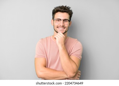 Young Handsome Man Looking Happy And Smiling With Hand On Chin, Wondering Or Asking A Question, Comparing Options