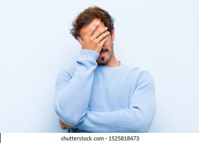 young handsome man looking stressed, ashamed or upset, with a headache, covering face with hand isolated against flat wall
