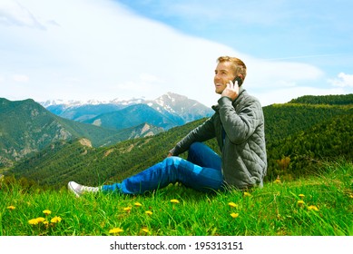 Young handsome man making a call on his mobile sitting in grass on green field. Casual wearing man talking on Cellphone outdoor. Mountains. Communication concept. Freedom. Vacation. Technology.