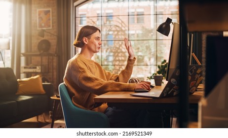 Young Handsome Man Making a Video Call on Desktop Computer in Sunny Stylish Loft Apartment. Creative Person Wearing Cozy Yellow Sweater and Headphones. Urban City View from Big Window.