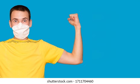 Young Handsome Man In Medical Mask And Yellow T-shirt Isolated On Blue Background. Strong Person Showing Arm Muscle, Confident And Proud Of Power To Dispose Of Problem