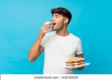 Young handsome man over isolated blue background holding mini cakes and eating it