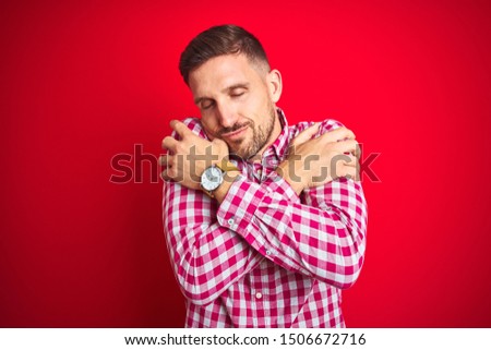 Young handsome man over red isolated background Hugging oneself happy and positive, smiling confident. Self love and self care