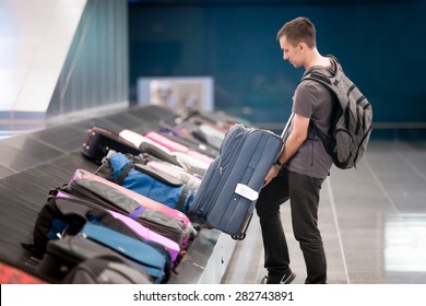 Young Handsome Man Passenger In 20s With Carry-on Backpack Collecting His Luggage At Conveyor Belt In Arrivals Lounge Of Airport Terminal Building