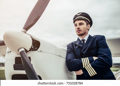 Young Handsome Man Pilot In Uniform Is Standing Near Small Private Plane.