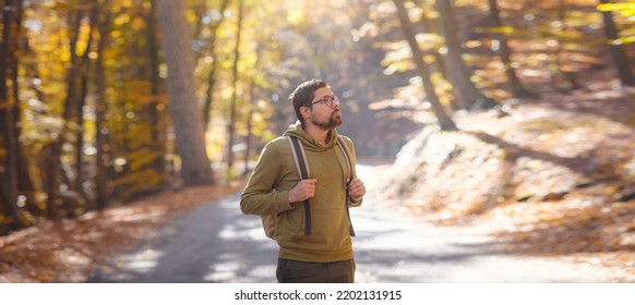 Young Handsome Man Posing In Autumn Forest. Young Hipster Guy With Backpack , Traveller Standing In Woods, Hiking, Forest, Journey, Active Healthy Lifestyle, Adventure, Vacation Concept.