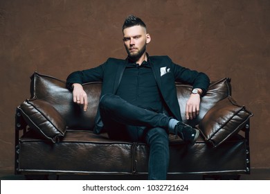 Young Handsome Man Posing For A Fashion Shoot In A Studio. Fashion As A Lifestyle. Man Wearing A Beard. Model Sitting On A Sofa In Tuxedo. Successful Fashionable Businessman. Business Look Advertising