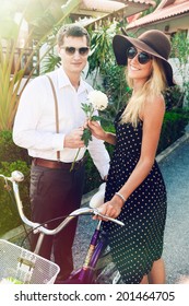 Young Handsome Man Present Flower To His Girlfriend At First Date. Posing Outdoor In City Garden With Retro Bike, Wearing Stylish Vintage Outfit And Sunglasses.