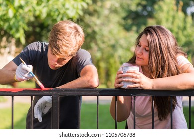 Young Handsome Man And Pretty Girl Are  Painting Metal Fence With A Brush