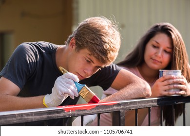 Young Handsome Man And Pretty Girl Are  Painting Metal Fence With A Brush