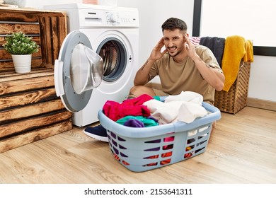 Young Handsome Man Putting Dirty Laundry Into Washing Machine Covering Ears With Fingers With Annoyed Expression For The Noise Of Loud Music. Deaf Concept. 