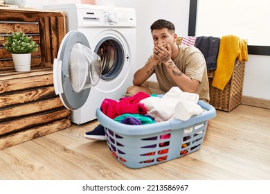 Young Handsome Man Putting Dirty Laundry Into Washing Machine Shocked Covering Mouth With Hands For Mistake. Secret Concept. 