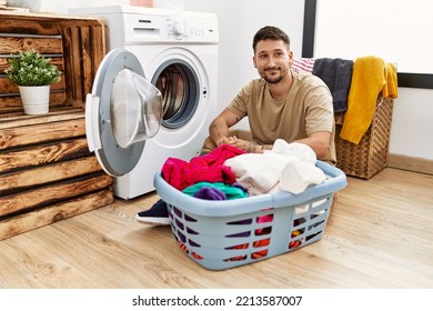 Young Handsome Man Putting Dirty Laundry Into Washing Machine Smiling Looking To The Side And Staring Away Thinking. 