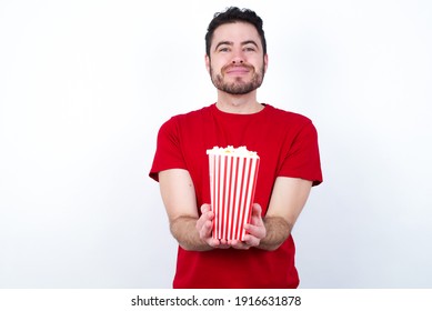 Young handsome man in red T-shirt against white background eating popcorn holding something with open palms, offering to the camera.