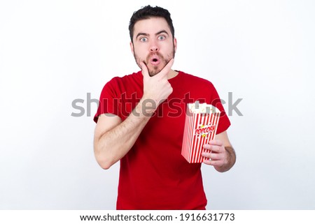 Young handsome man in red T-shirt against white background eating popcorn Looking fascinated with disbelief, surprise and amazed expression with hands on chin