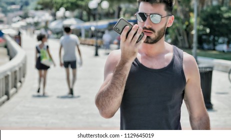 Young handsome man at the seaside using smartphone to record and send voice message, while looking at the beach and the sea