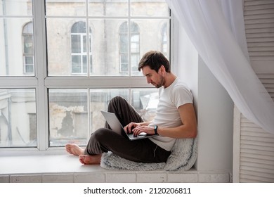 A Young Handsome Man Sits On A Wooden Windowsill Near The Window In A Lotus Position, Works On A Gray Laptop. Remote Work Freelancer. The Programmer, Hacker Is Typing On A Computer At Home In Pajamas