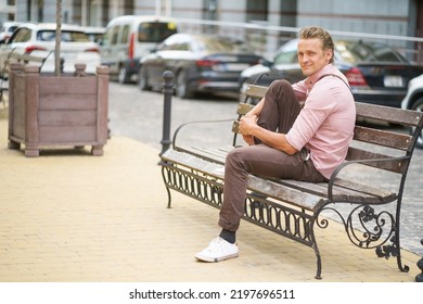 Young Handsome Man Sits On The Bench With One Leg Up On It Spend Time In Urban City Near His Office Or Home, Wearing Pink Shirt And Shoulder Bag. Freelancer Man Off Work. 