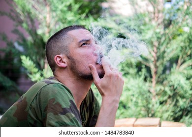 Young Handsome Man Is Sitting In The Backyard Of His House And Smoking A Weed. War Veteran Enjoys In Marijuana Joint In The Garden.