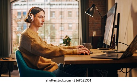 Young Handsome Man Sitting Down to Work on Desktop Computer in Sunny Stylish Loft Apartment. Creative Designer Wearing Cozy Yellow Sweater and Headphones. Urban City View from Big Window.