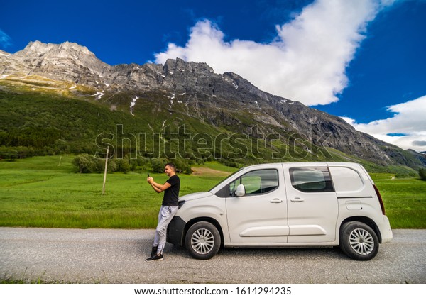 Young\
handsome man sitting near hired car and use the mobile phone on the\
norway mountains background. Car hire. Car\
trip.