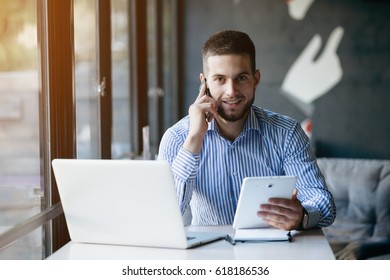 Young Handsome Man Sitting In Office With Cup Of Coffee And Working On Project Connected With Modern Cyber Technologies. Businessman With Notebook Trying To Keep Deadline In Digital Marketing Sphere.