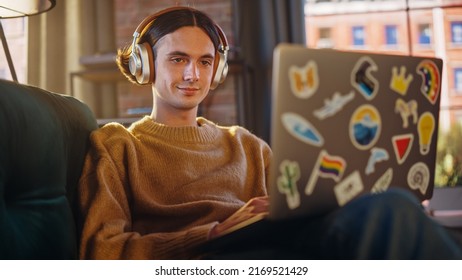 Young Handsome Man Sitting on Sofa and Using Laptop Computer in Sunny Stylish Loft Apartment. Creative Designer Wearing Cozy Yellow Sweater and Headphones. Urban City View from Big Window.