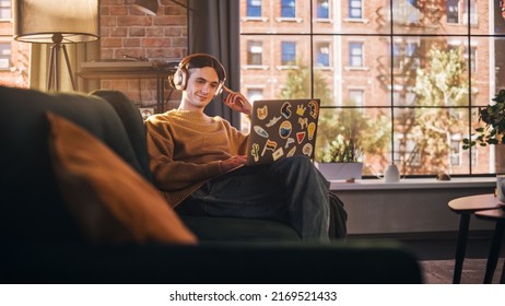 Young Handsome Man Sitting on Sofa Works on Laptop Computer in Sunny Stylish Loft Apartment. Creative Designer Wearing Cozy Yellow Sweater and Headphones. Urban City View from Big Window.
