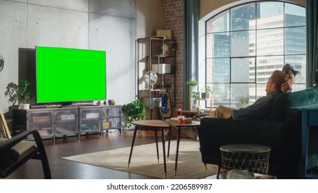 Young Handsome Man Sitting On A Sofa And Watching TV With Horizontal Green Screen Mock Up. It's Day Time On Weekend At Home. Spacious Living Room In Stylish Loft Apartment.