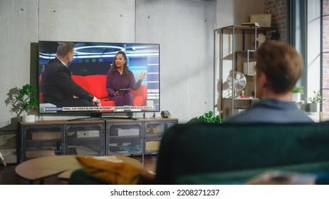 Young Handsome Man Sitting In Relaxed Pose On A Sofa And Watching TV With Live Talk Show During The Day Time On Weekend. Serious Female African American Host Interviews Guest In Studio.