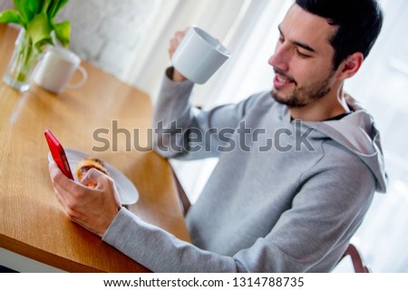 Young handsome man sitting at table with cup of coffee or tea and mobile phone. Kitchen location in morning