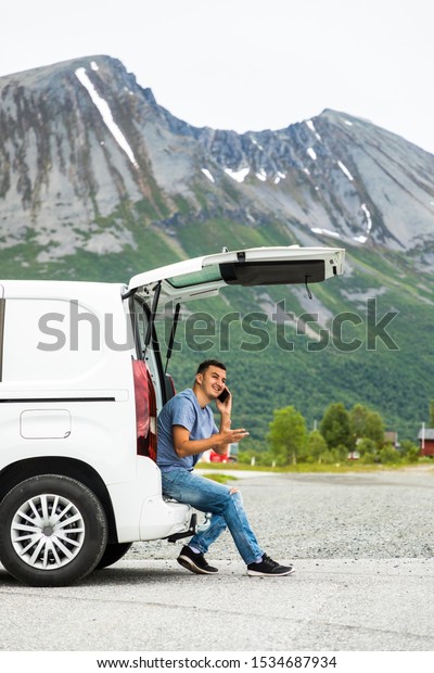 Young handsome man sitting in the trunk of his\
hired car and talking on the mobile phone on mountains background.\
Car hire. Car trip.