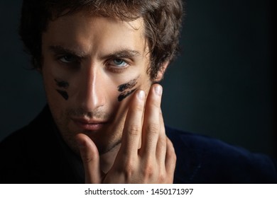 A Young Handsome Man With A Slight Unshaven Face And Decisiveness In His Eyes, Puts A Black War Paint On His Cheeks With His Fingers. Portrait On A Dark Background.