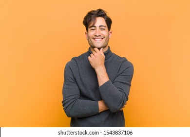 young handsome man smiling  enjoying life  feeling happy  friendly  satisfied   carefree and hand chin against orange background